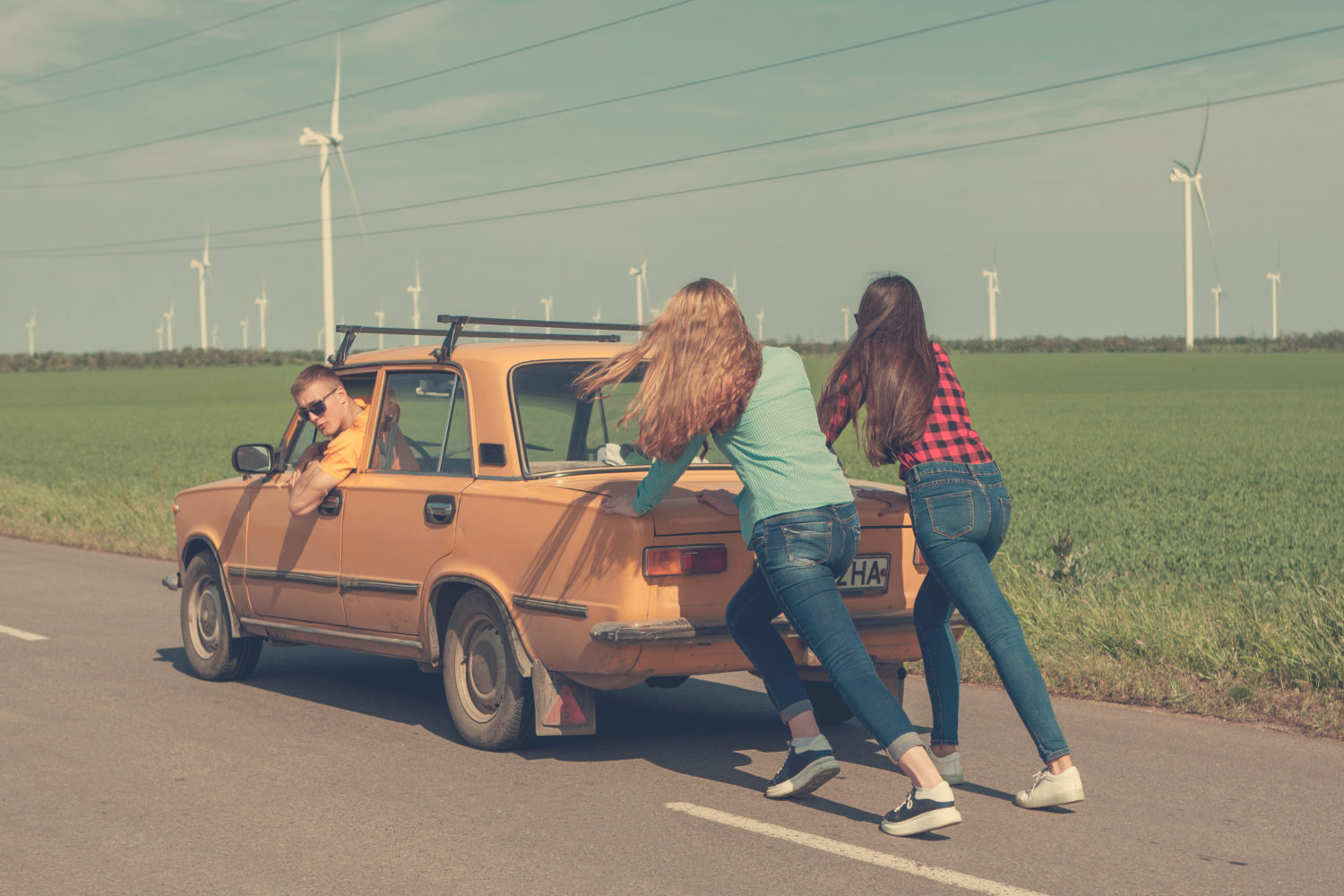 Two girl pushing an old orange Soviet car with a boy in it down the road while on a road trip from Riga to Vilnius