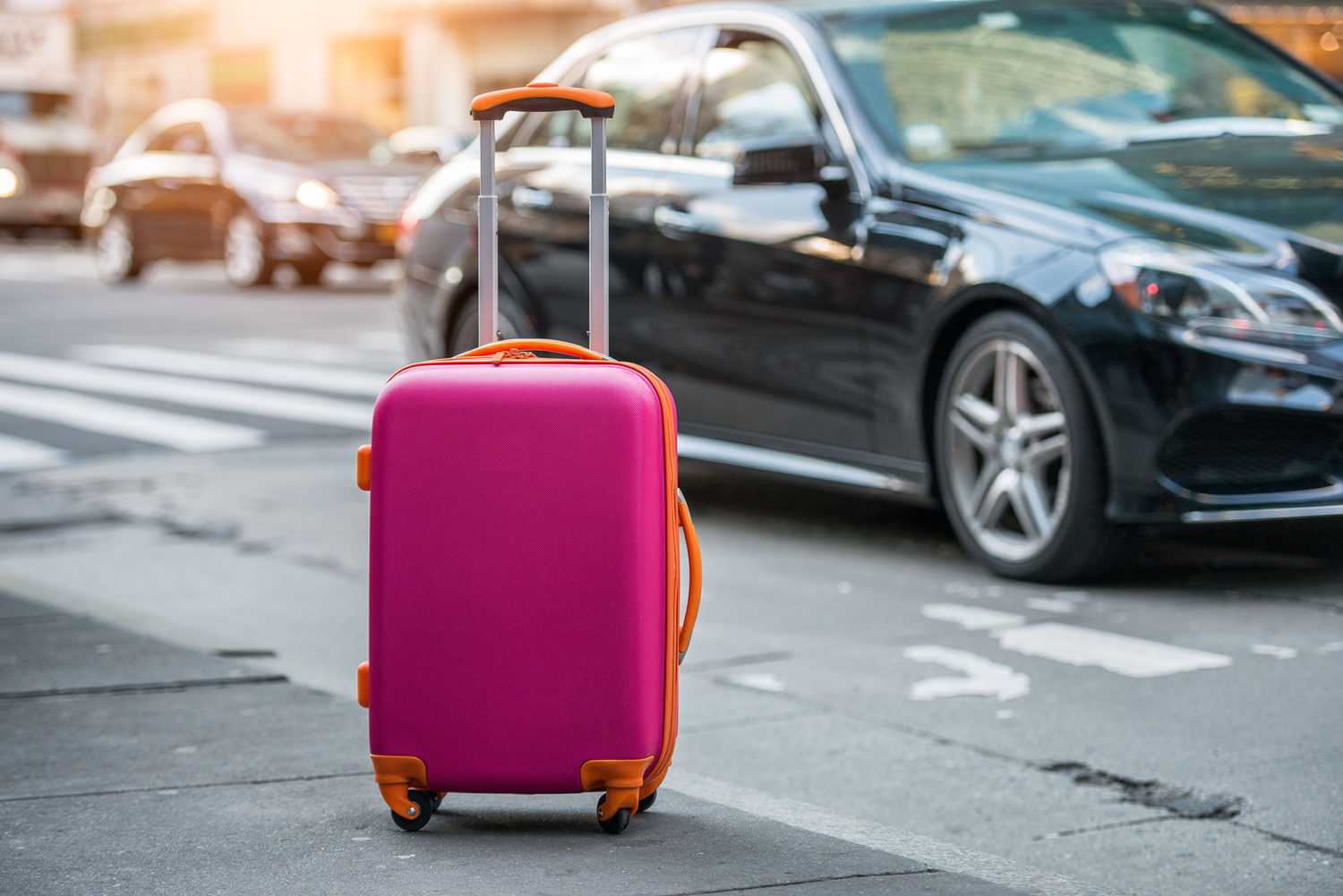 Pink suitcase waiting on a sidewalk with black luxury car in background
