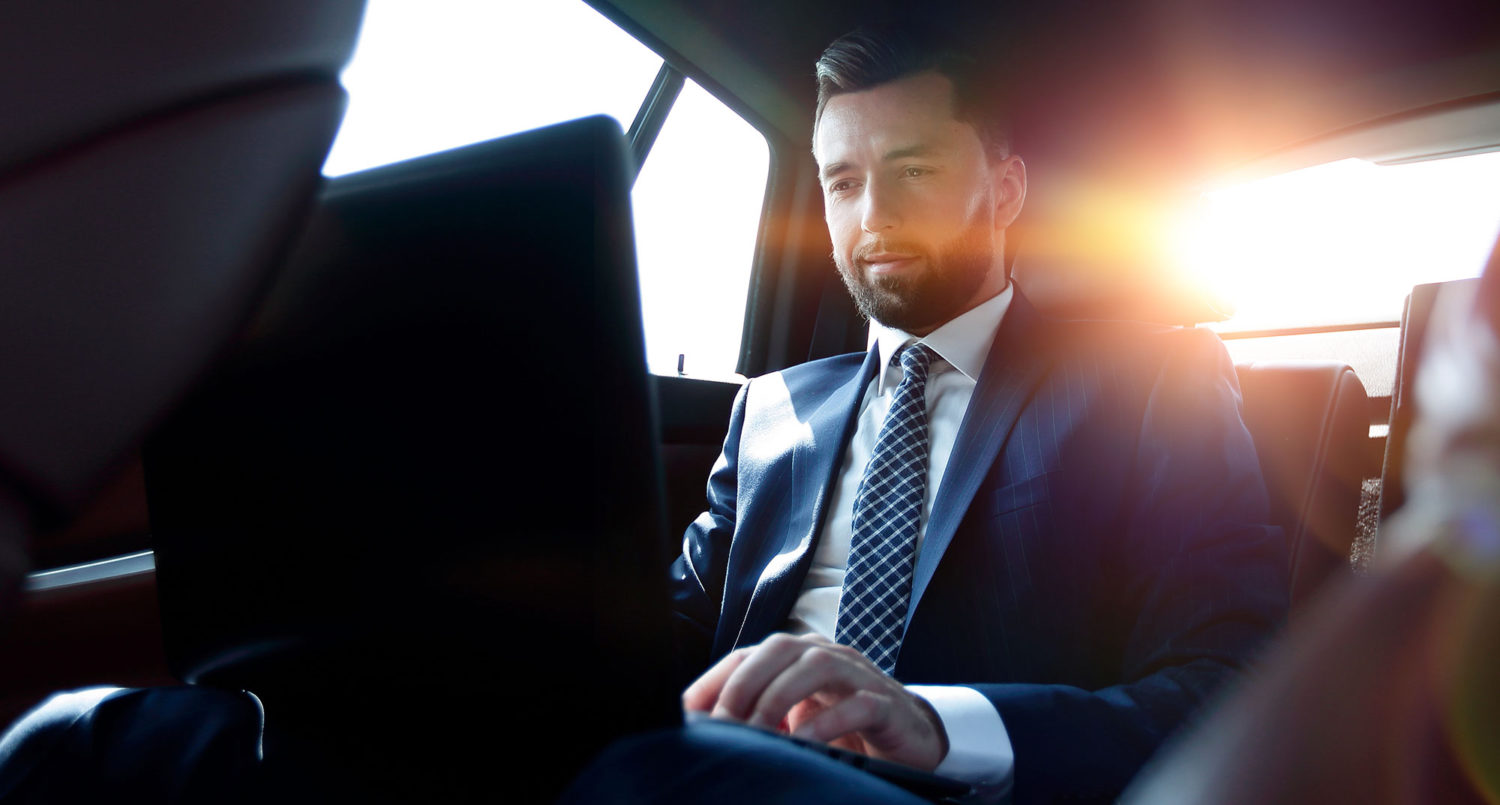 A man in a suit working with a laptop on the backseat of a car